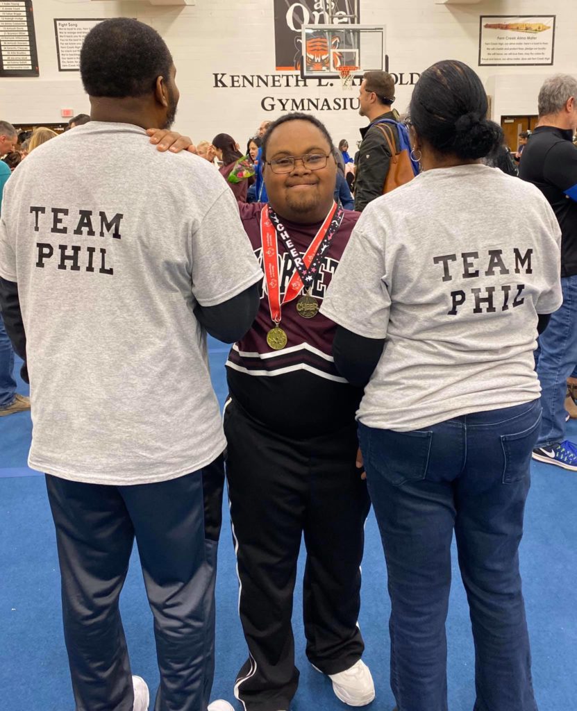 a young black man with down syndrome smiles proudly in a cheerleading uniform with a medal around his neck, flanked by two people wearing "team phil" t-shirts